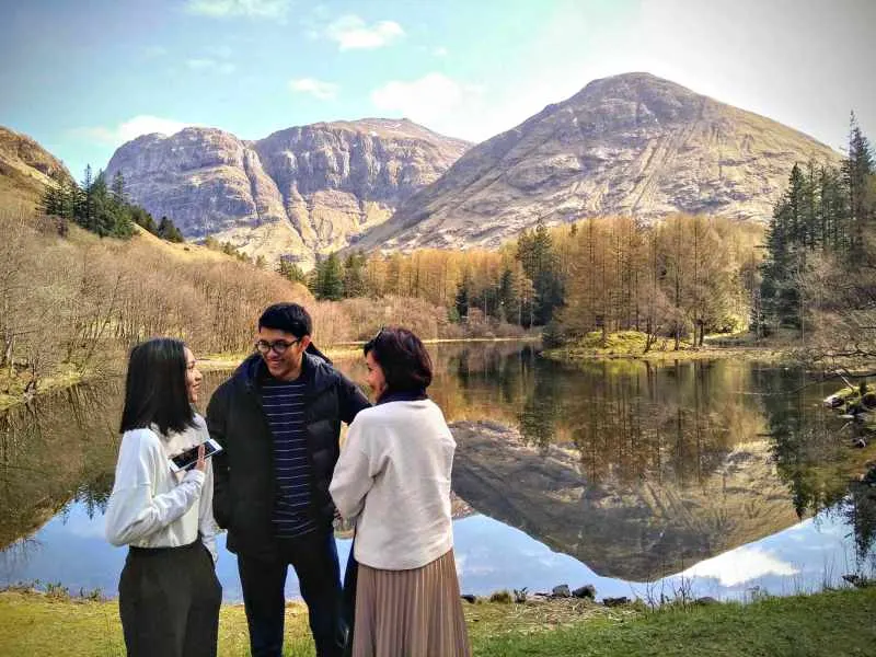Three people stand on a grassy riverbank facing each other, with mountains and their reflections in calm water behind them.