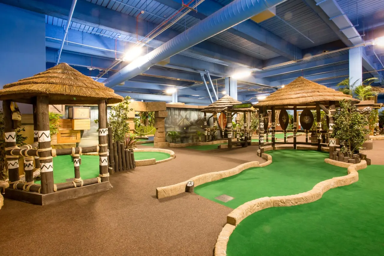Indoor miniature golf course with African-themed huts, green courses, and wooden decorative structures under industrial ceiling.