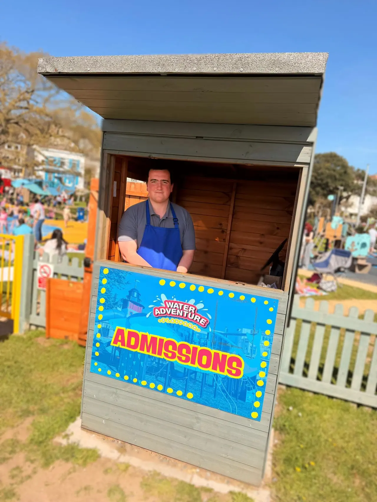Staff member at admissions booth with water park signage, busy outdoor leisure venue in background.