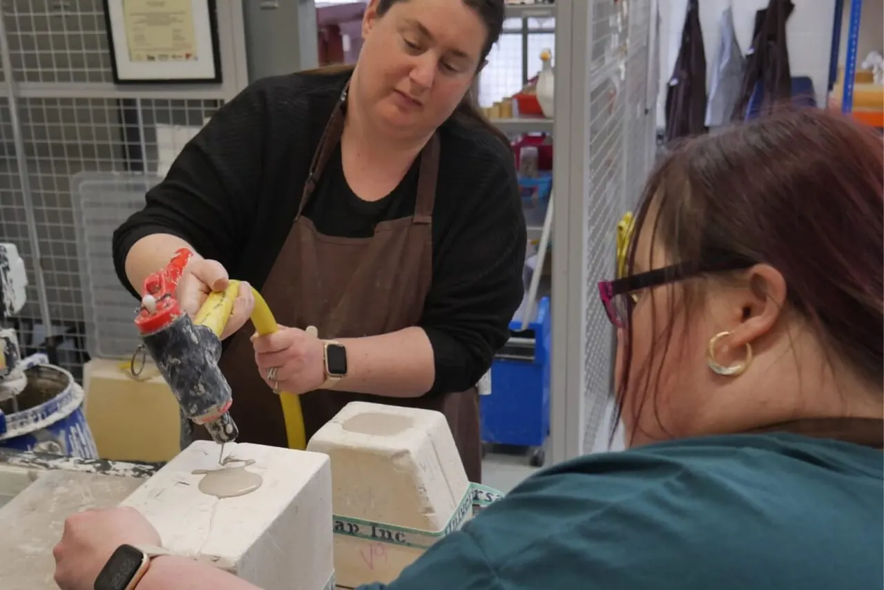 Woman in brown apron demonstrates glazing technique on a small ceramic piece to a child at a pottery workshop table.