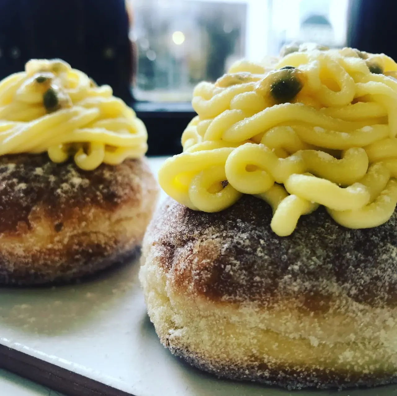 Two pastries with yellow icing piped on top and capers, dusted with sugar, served on a plate.