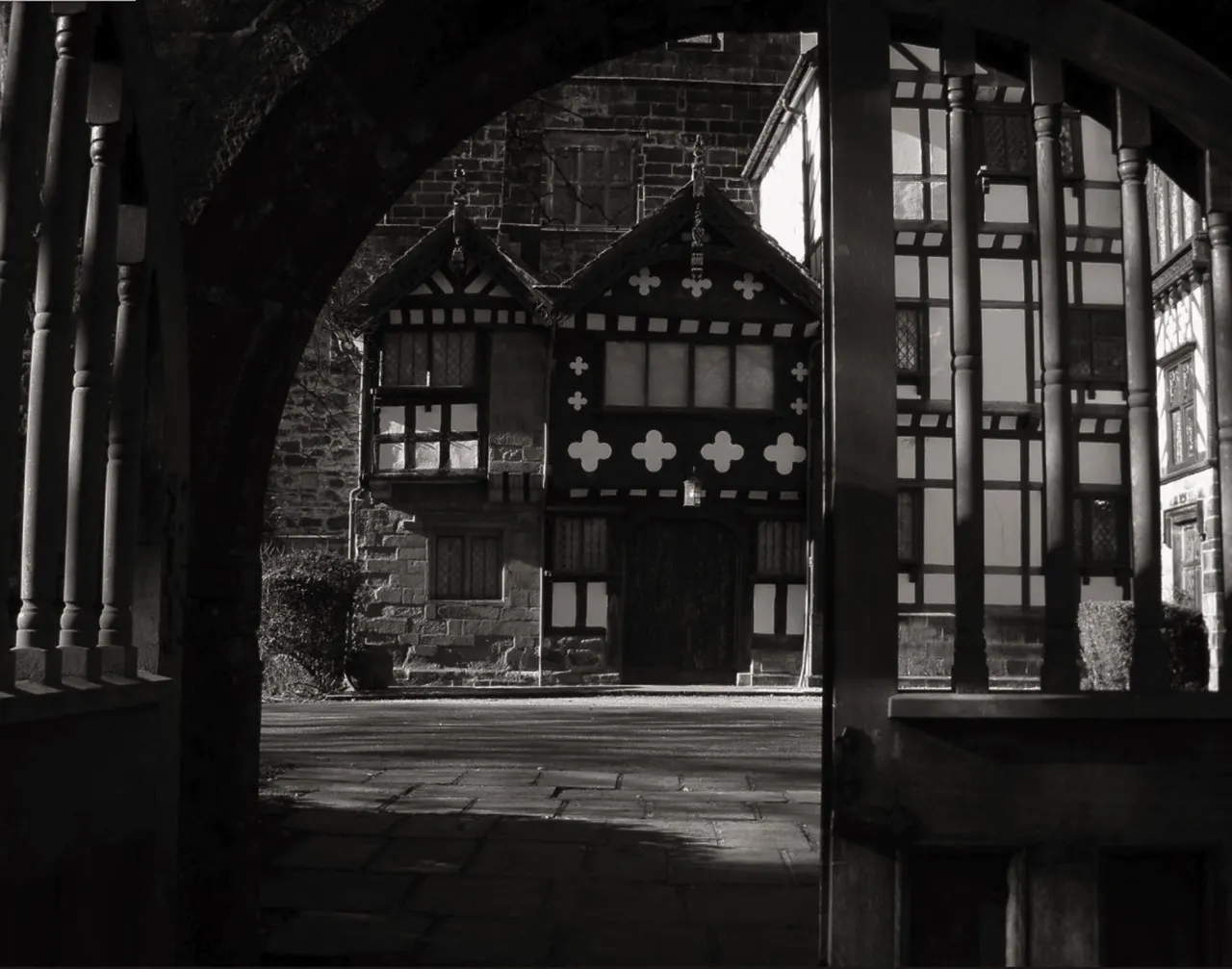 Black and white photograph of a Tudor-style building with decorative timber framing and cross motifs, viewed through a covered passageway.