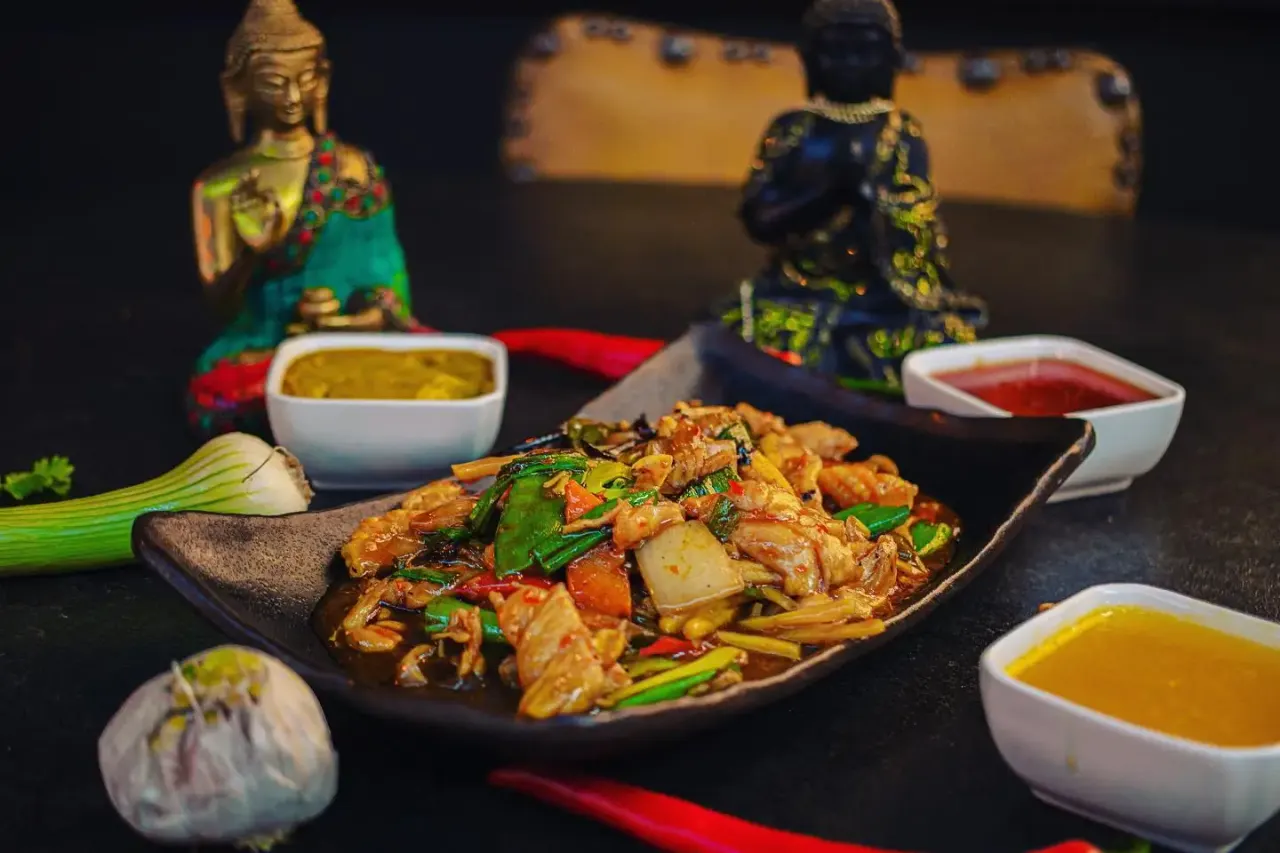 Plated Asian stir-fry dish with vegetables and sauce bowls, surrounded by decorative figurines and fresh ingredients.