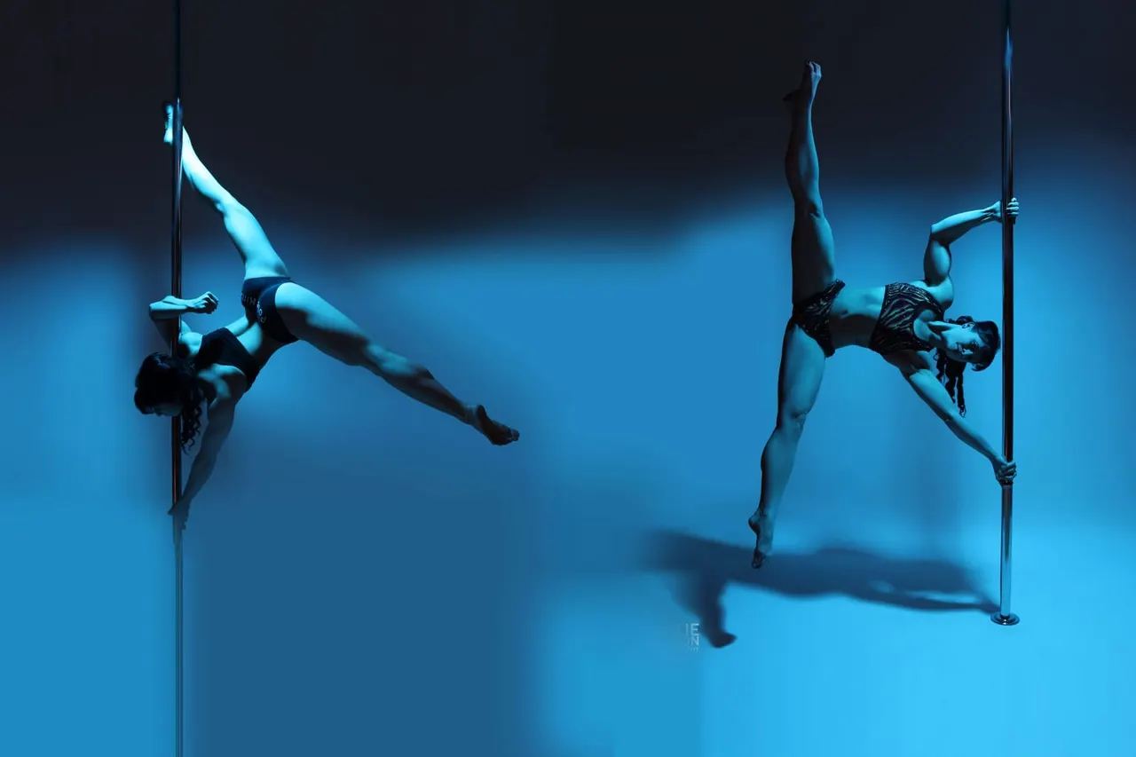 Two pole dancers performing aerial tricks on vertical poles against a blue-lit background.