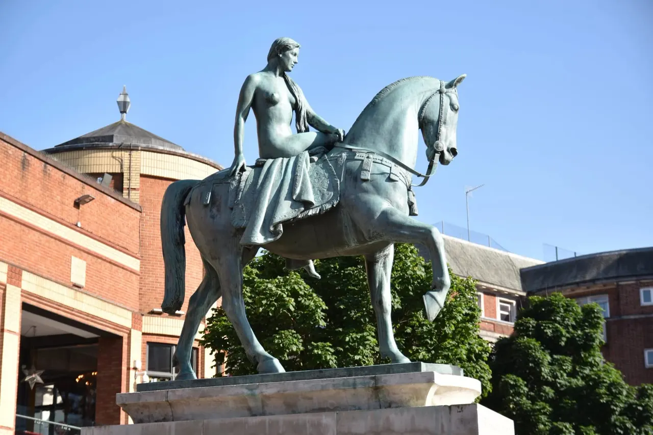 Bronze statue of a classical female figure riding a horse, displayed in a courtyard surrounded by brick buildings.