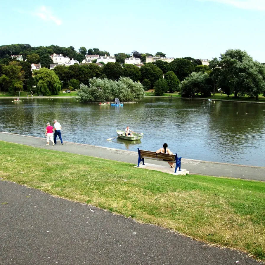 People enjoy a riverside park with rowing boats and waterside benches, Georgian villas visible on the hillside beyond.