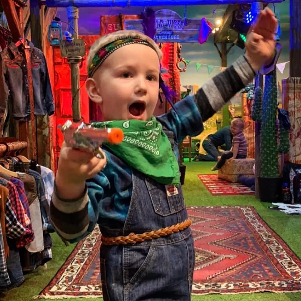 Young child in cowboy outfit enjoying an indoor western-themed play area with colourful neon lighting and props.