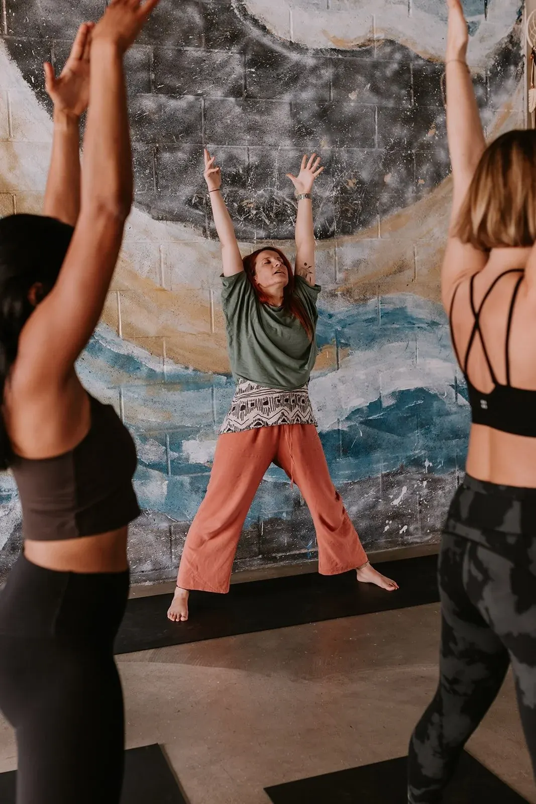 A woman jumps with arms raised during a fitness class whilst others exercise around her in front of a mural.