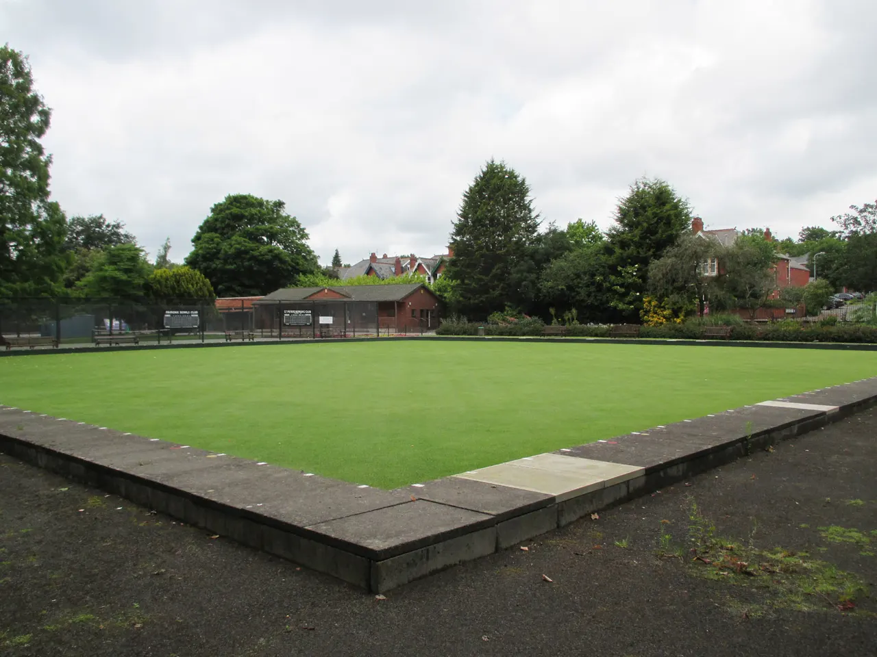Flat green lawn with a bowling green in the foreground, pavilion building and residential properties in the background.