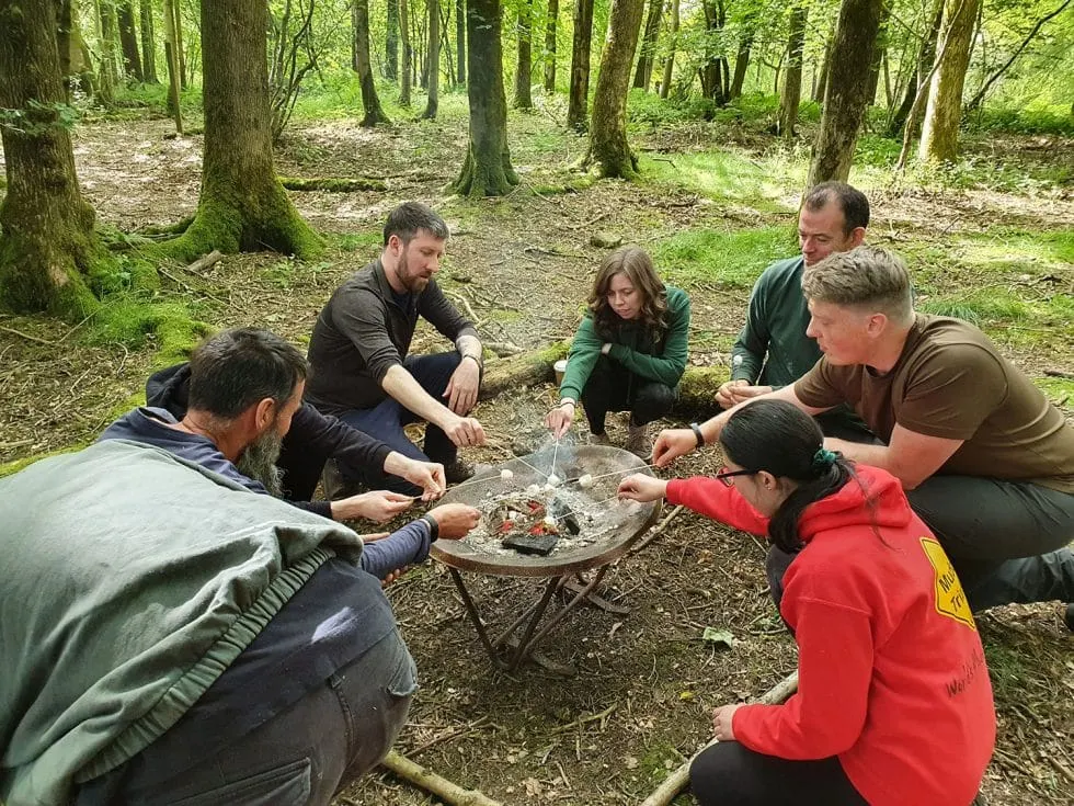 Group of six people gathered around a campfire in a woodland setting during an outdoor activity.