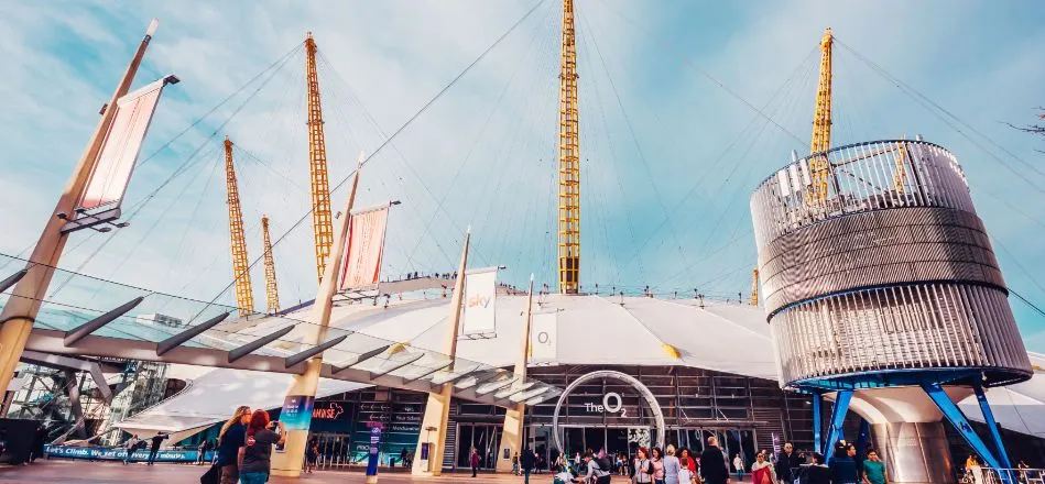 Crowds of visitors outside the distinctive white fabric roof and golden support towers of The O2 arena on a sunny day.