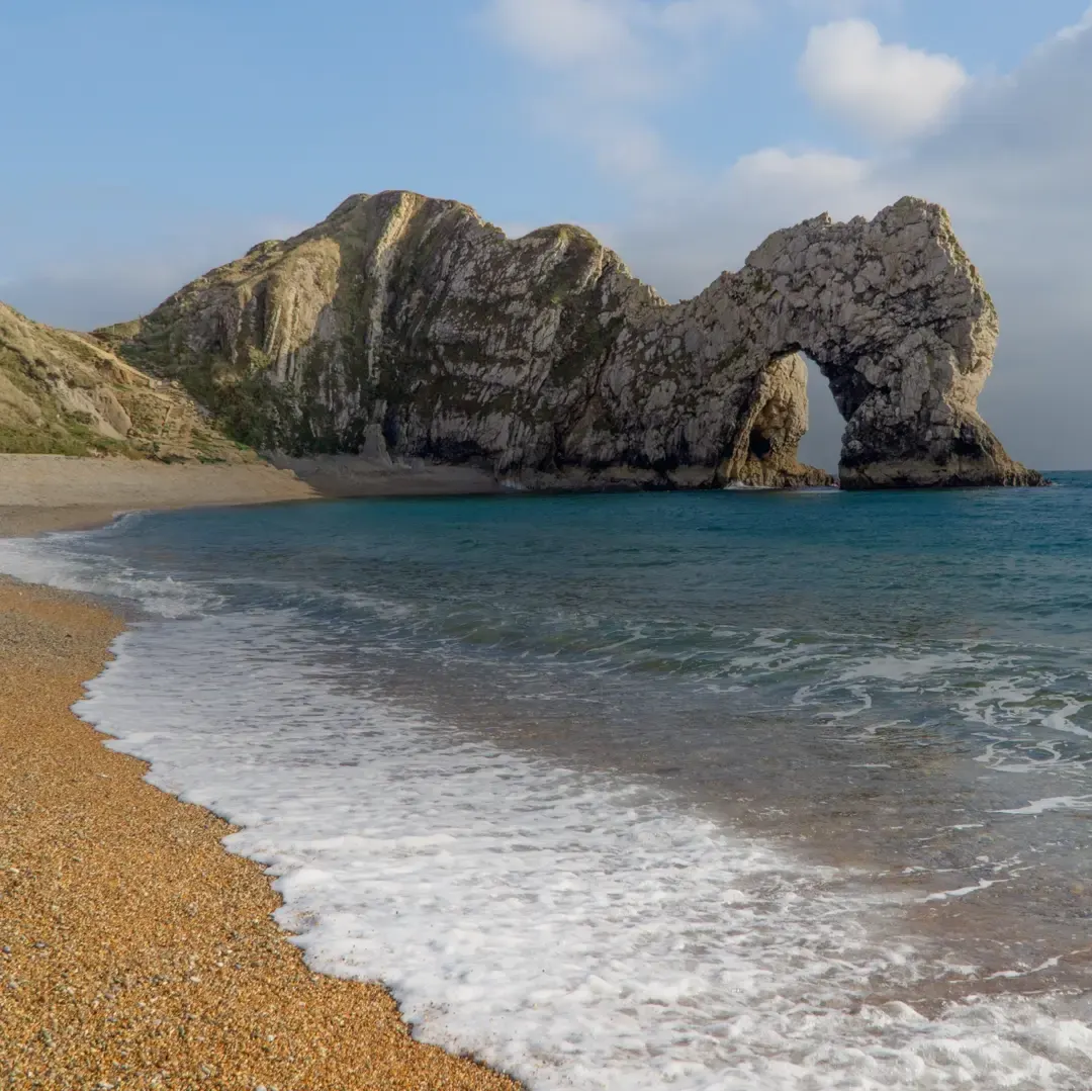 Rocky limestone arch formation rising from turquoise sea with sandy beach and gentle waves in foreground.