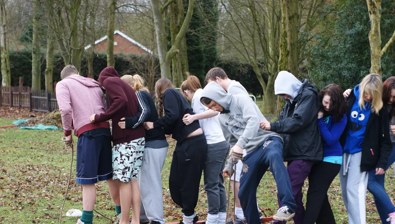 Group of young people gathering around an outdoor activity in a wooded area with bare trees and a building visible in the background.
