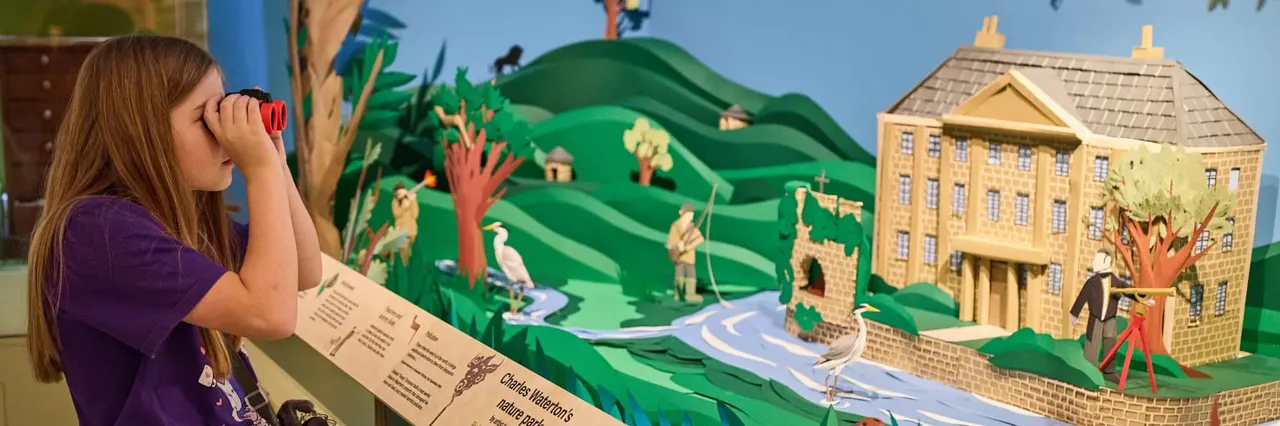 Child holding red binoculars whilst viewing a diorama display of a yellow stone building and green landscape.