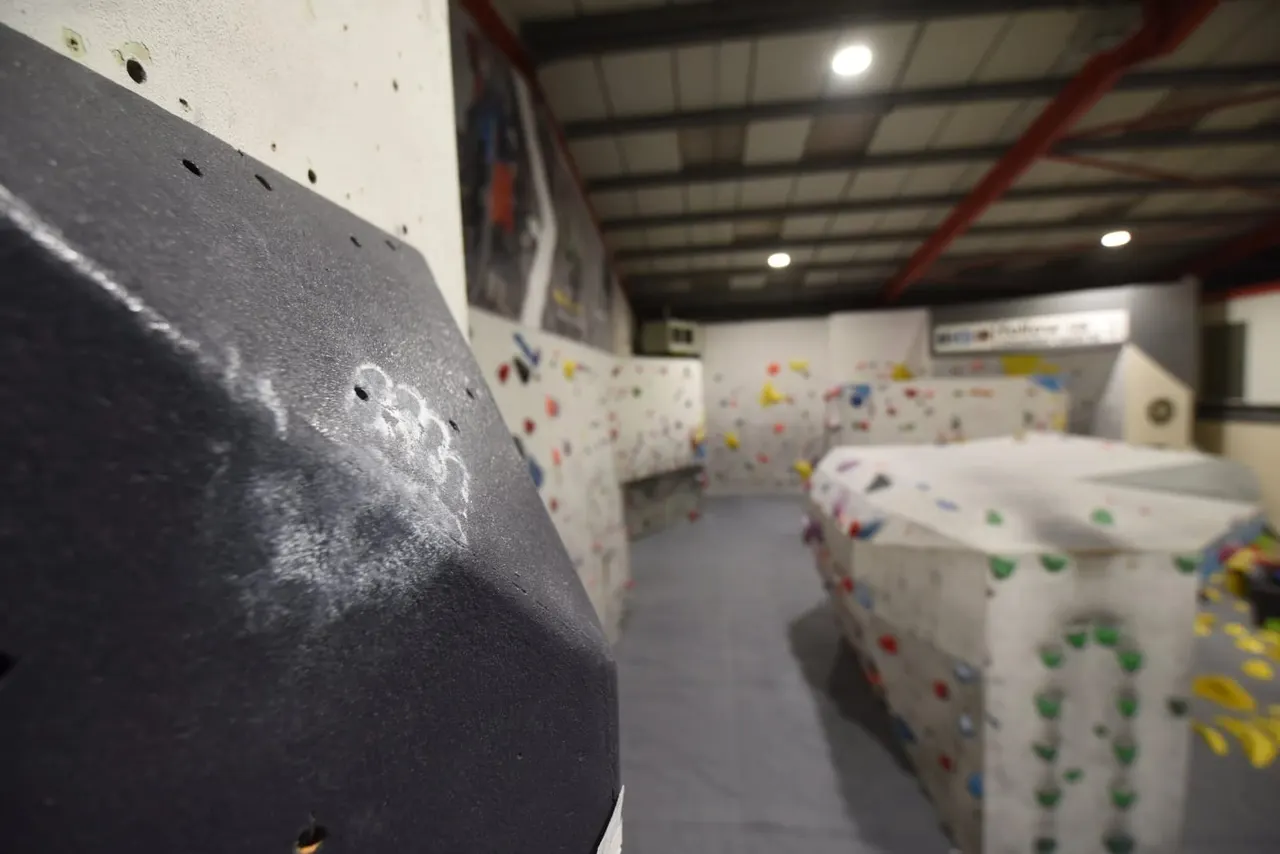 Interior of an indoor climbing facility with multiple white climbing walls featuring coloured handholds, and a dark padded wall panel in the foreground.