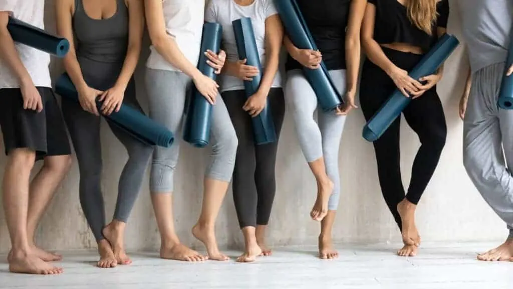 Group of barefoot people in fitness wear holding rolled yoga mats, standing together against a white wall.