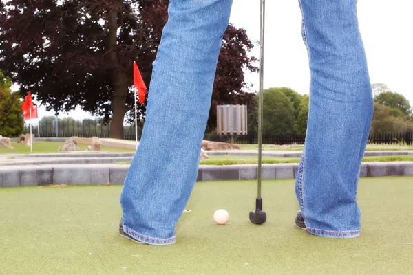 Person in jeans and red shoes putting on a mini golf green with a golf ball and club visible.