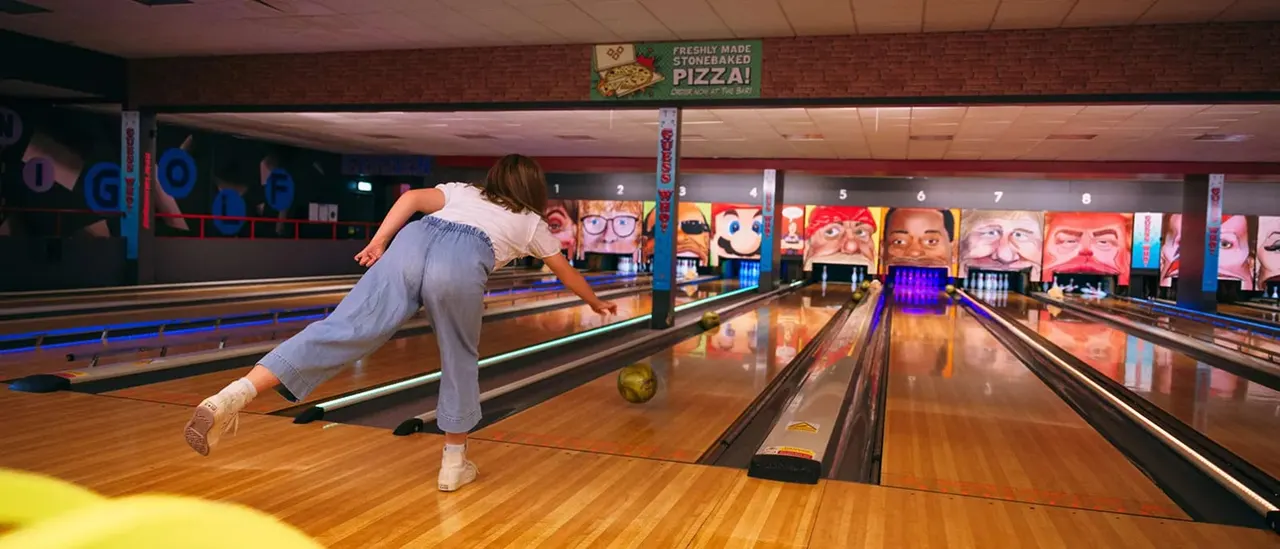 Woman bowling mid-release on a wooden lane in an indoor bowling alley with colourful face murals on the back wall.