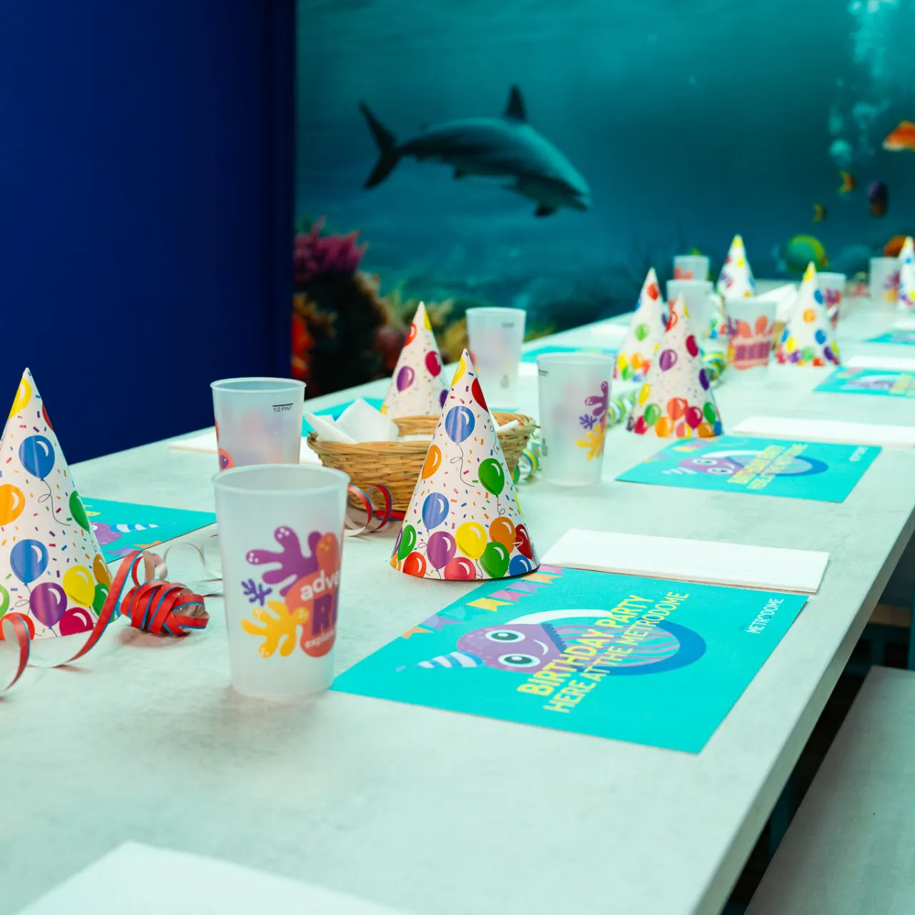Children's birthday party table set with colourful party hats, cups and placemats, overlooking an underwater aquarium tank with a shark and coral.