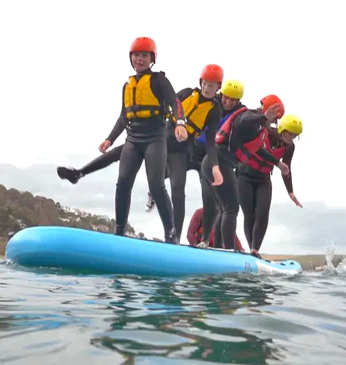 Group of people in wetsuits and helmets standing on a blue paddleboard in water.