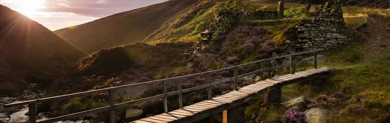 Wooden boardwalk bridge crossing moorland with stone ruins, rolling hills and heather at sunset.