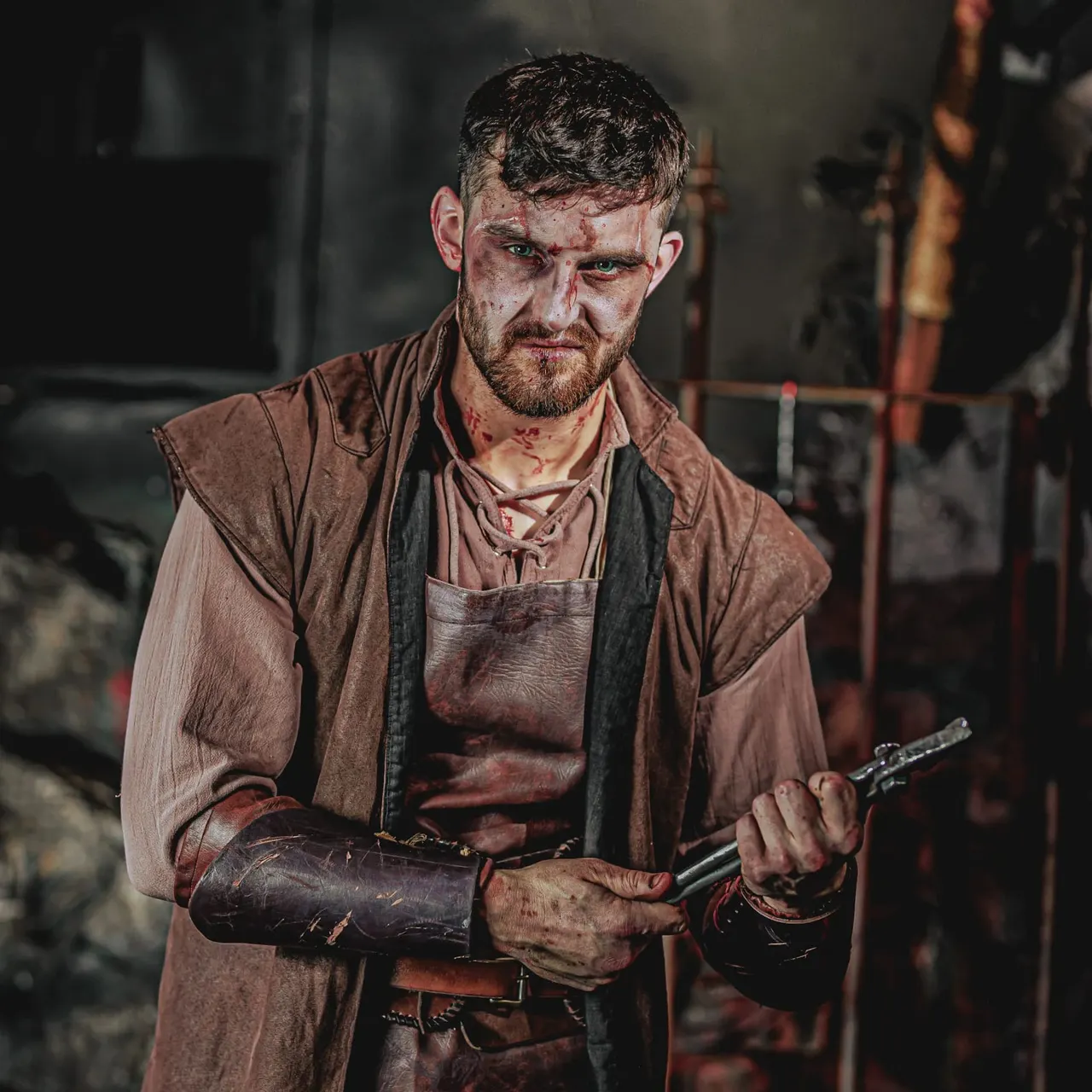 Actor in period costume with fake blood holding a weapon in a dark dungeon chamber.