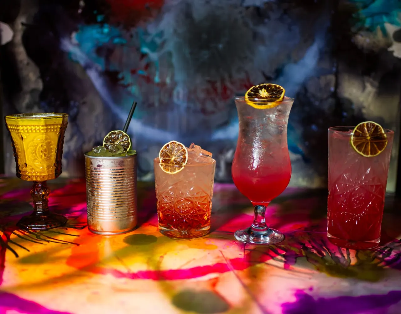 Five colourful cocktails with citrus garnishes arranged on a surface lit by vibrant multicoloured lighting.