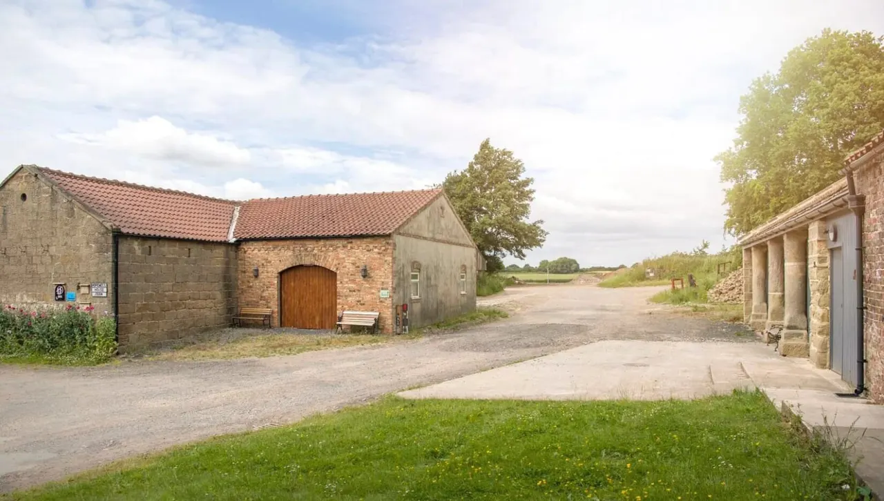 A rural farm courtyard with stone buildings and a gravel drive, surrounded by open countryside and trees.