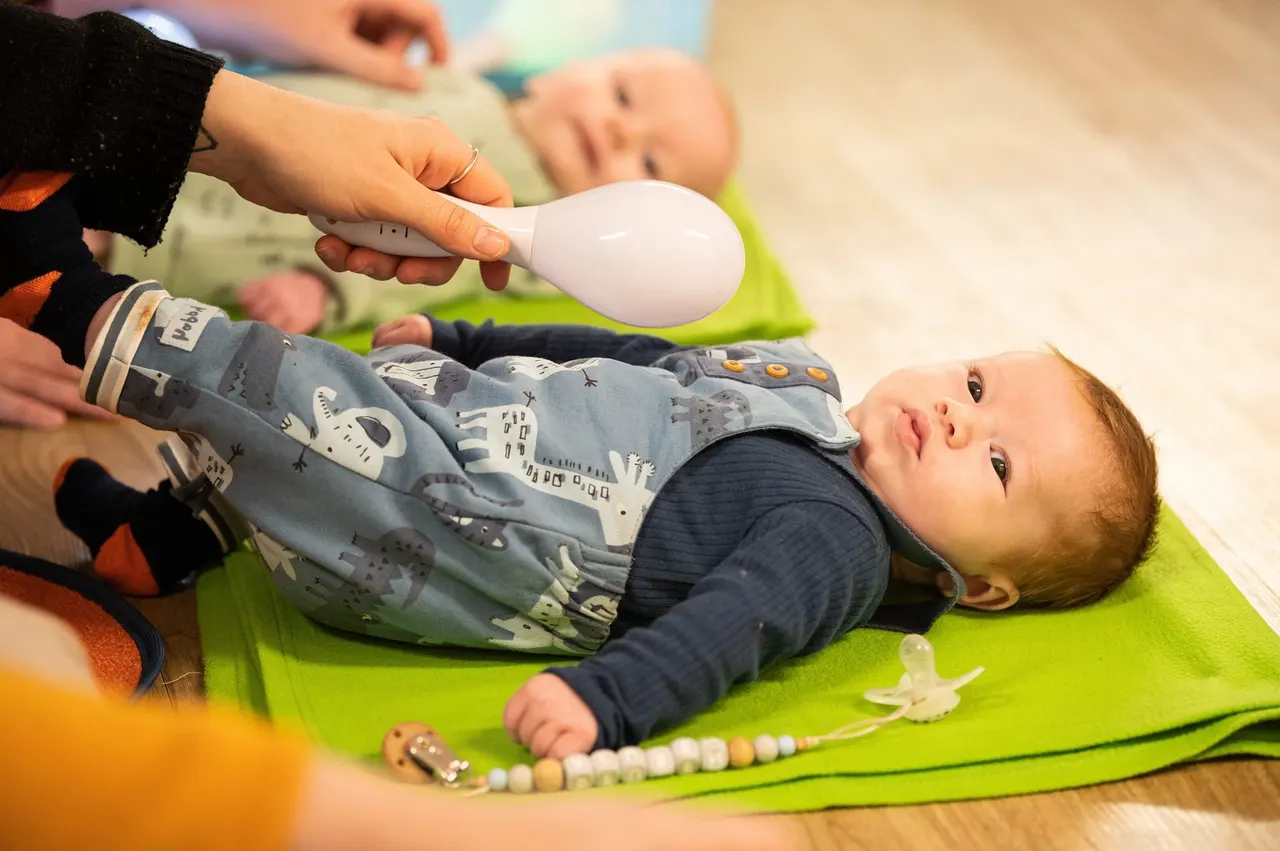 Infant on a bright green mat during a music class, being shown a white instrument by an adult's hand.
