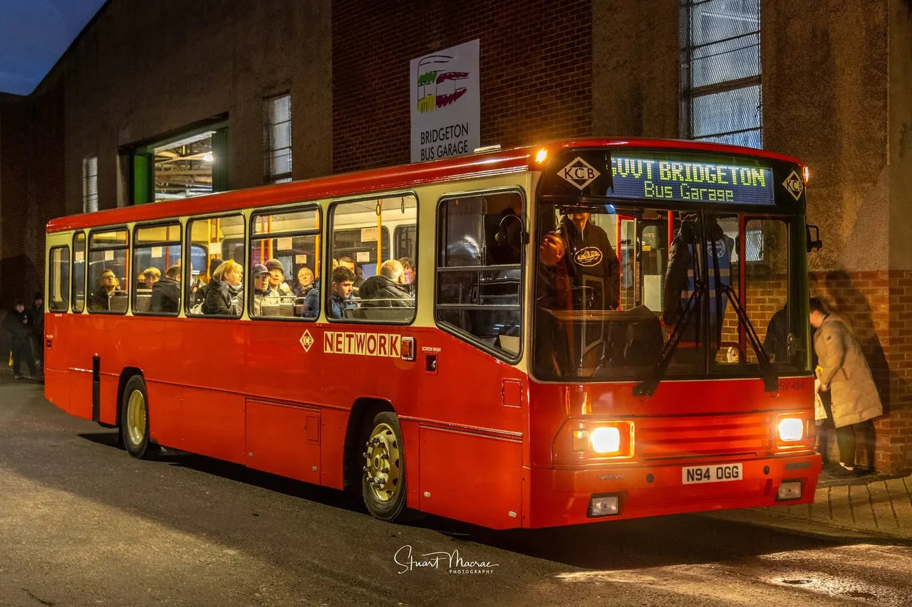 A red vintage bus with passengers visible through illuminated windows, parked at night outside a brick building with "Bridgeton Bus Garage" signage.
