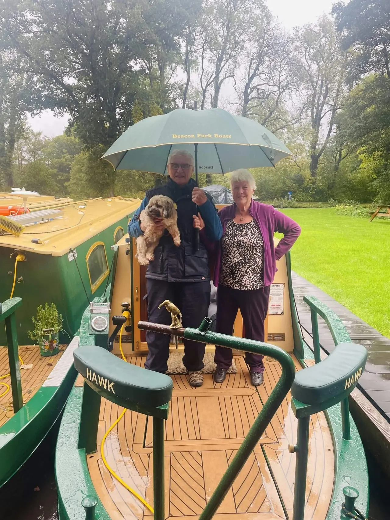 Two people and a dog stand on a narrowboat deck under an umbrella during rainy weather.