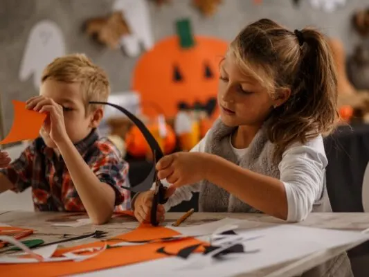 Two children at a table doing Halloween crafts with orange paper and decorations.