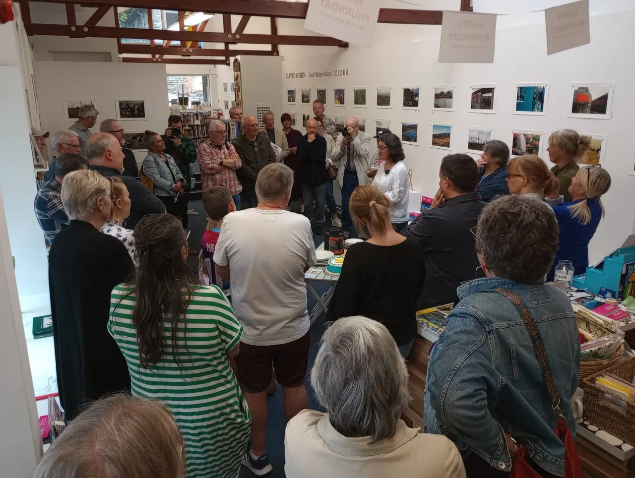A group of visitors gathered inside a gallery space listening to a speaker, with framed photographs displayed on the walls behind them.