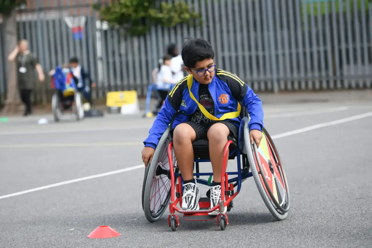 Young person in blue and yellow sports jacket racing a red wheelchair on an outdoor track with other participants in the background.