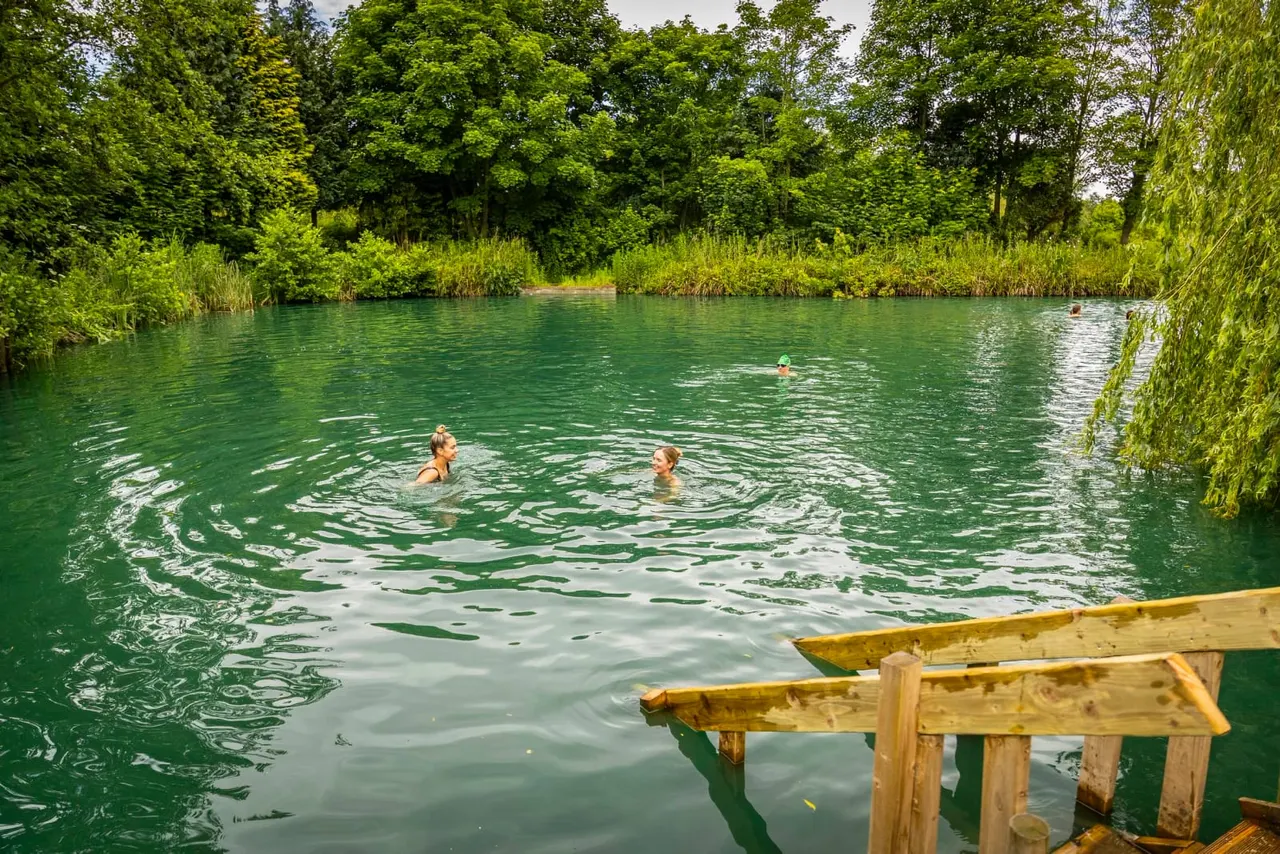 People swimming in an emerald-green river surrounded by lush trees and reeds, viewed from a wooden jetty.