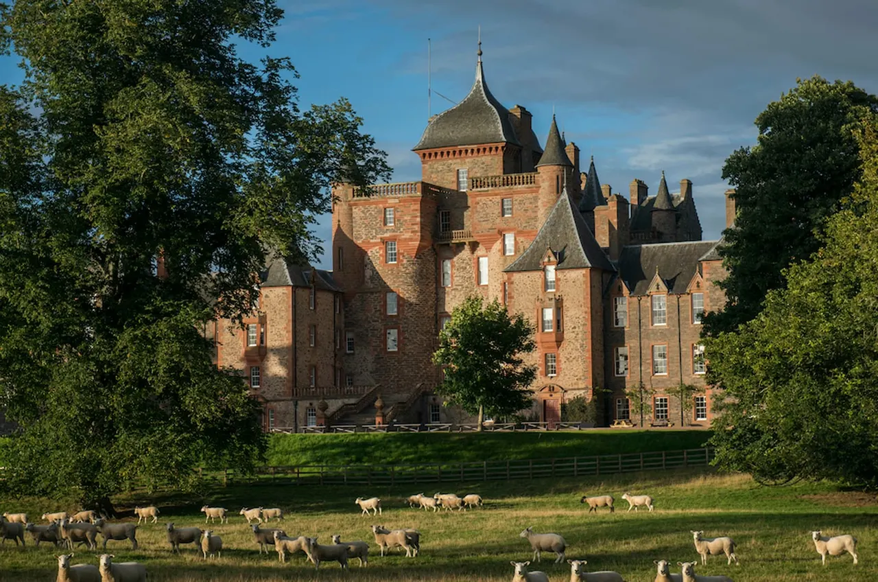 A red sandstone castle with multiple turrets and towers surrounded by trees, with a herd of sheep grazing in the parkland in front.