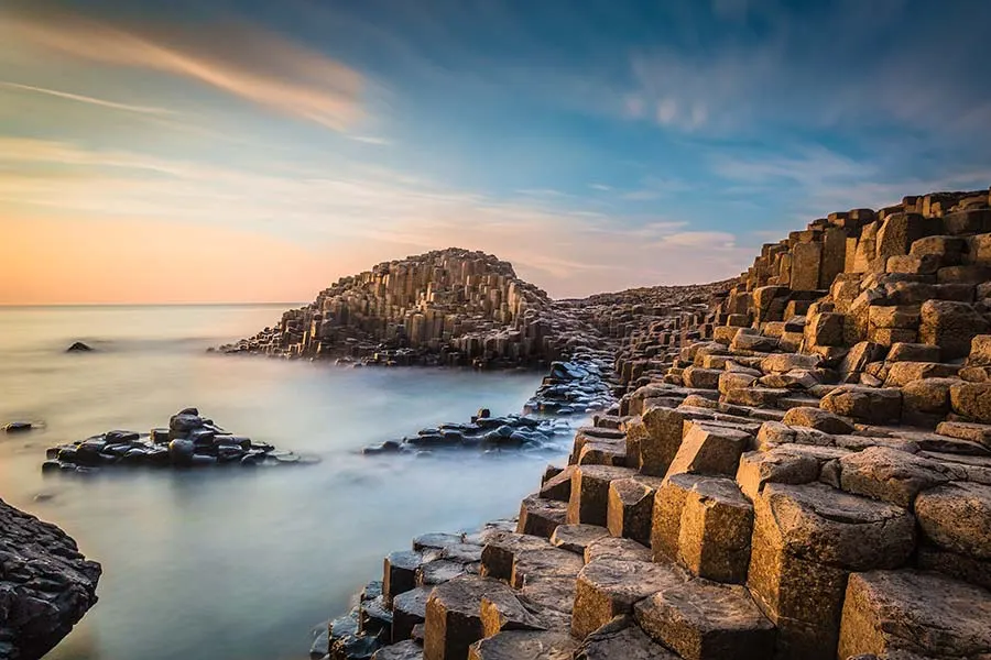 Dramatic coastal landscape with distinctive hexagonal rock columns extending into calm sea at sunset.
