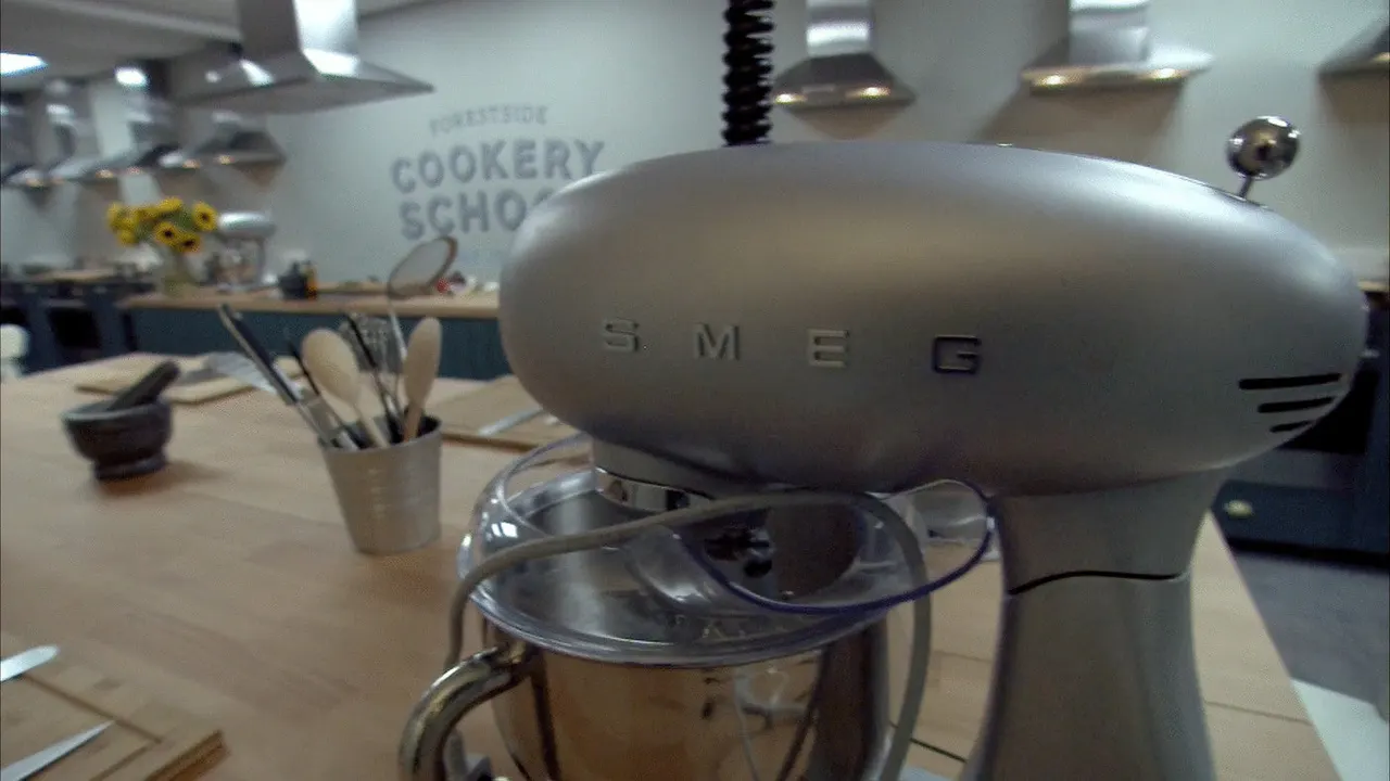 A silver SMEG food mixer sits on a wooden counter in a cookery school kitchen, with utensils and a blender nearby.