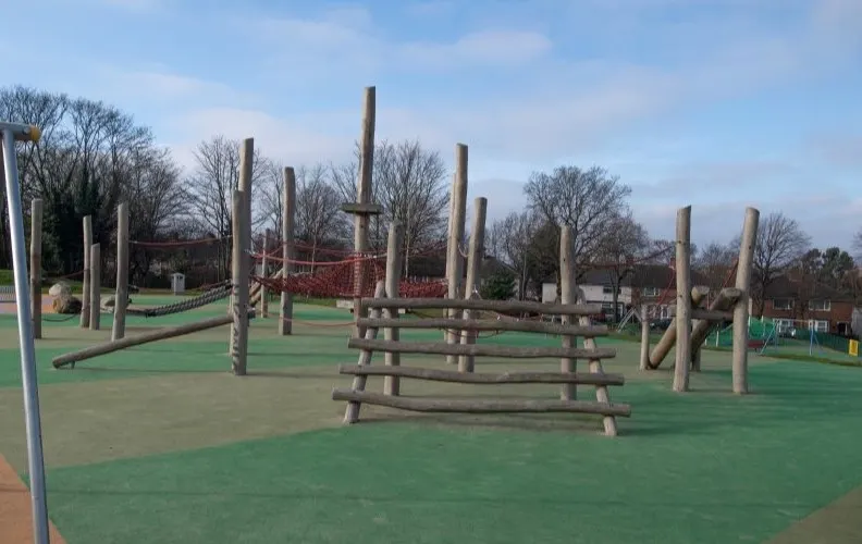 Wooden climbing and balance equipment on a green recreational playing surface in a public park.