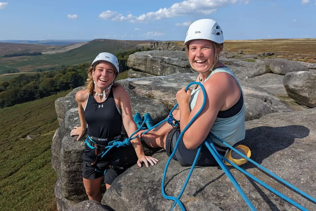Two women in safety helmets with climbing rope on a rocky outcrop overlooking moorland and valleys.