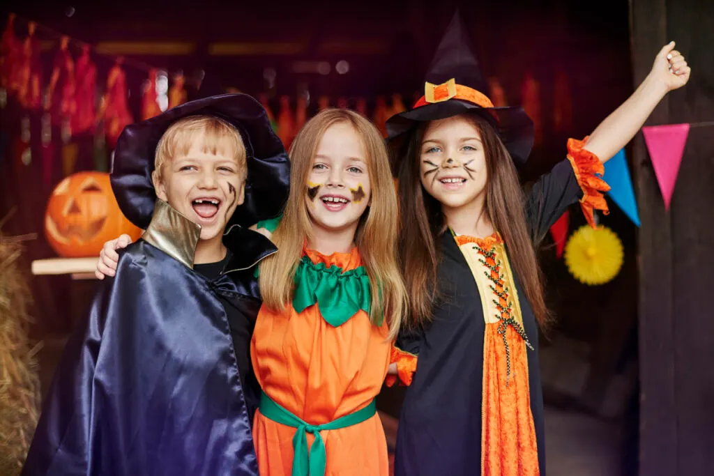 Three children in Halloween costumes smile at the camera in a decorated venue with pumpkins and colourful bunting.