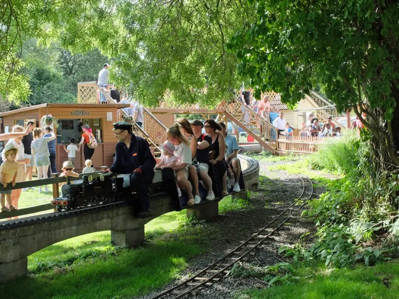 Passengers boarding a miniature steam train at a heritage railway attraction, with wooden structures and lush green foliage overhead.