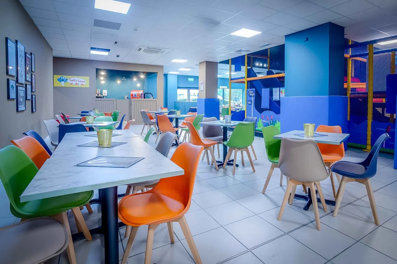 Bright indoor play facility with colourful chairs and tables, a soft play structure with yellow poles, and a service counter in the background.