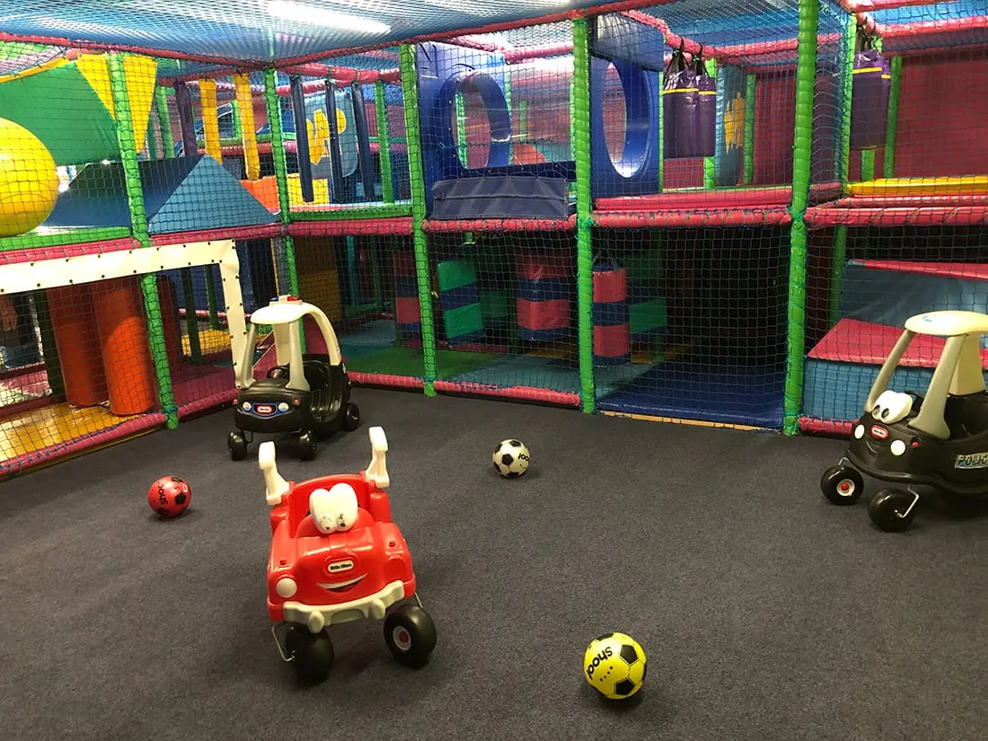 Colourful indoor play area with toy cars and footballs on a black floor, surrounded by netting and climbing structures.