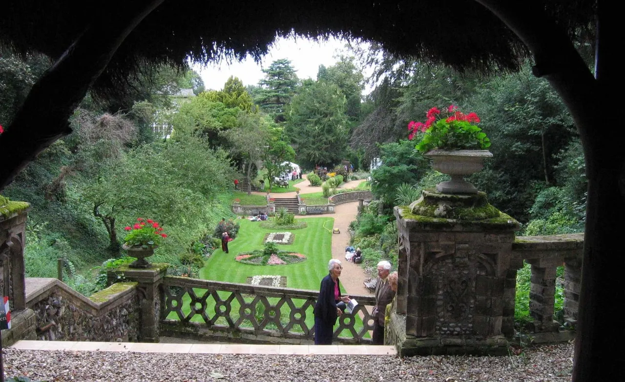 Visitors enjoying a formal walled garden with manicured lawns, stone balustrade, geranium-filled urns and mature trees.