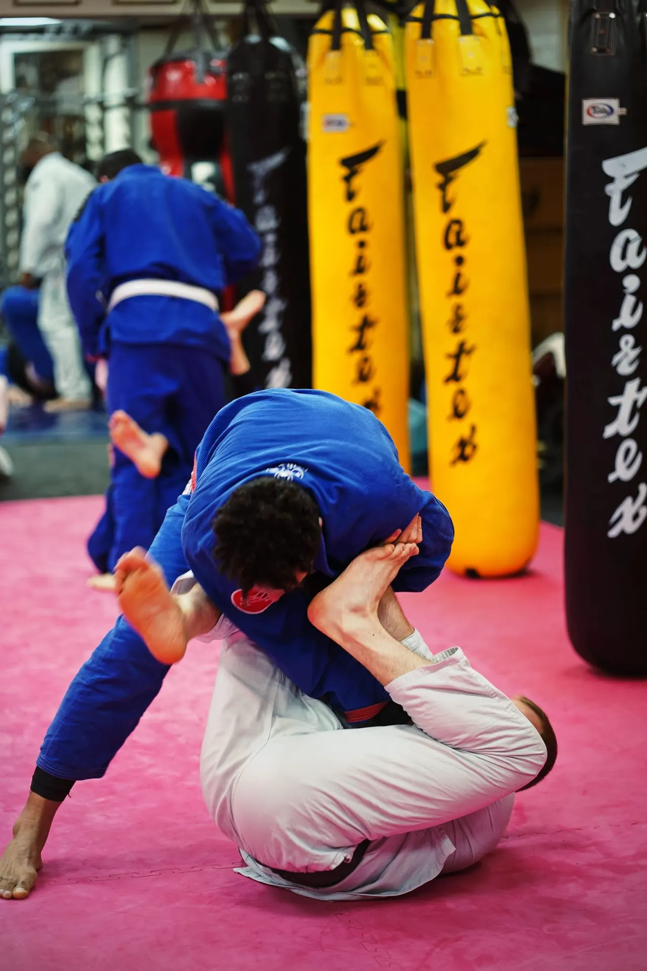 Martial arts student in blue gi performing a grappling technique on pink mat with heavy bags in background.