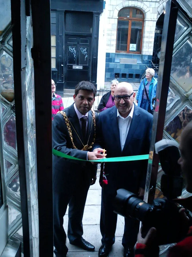 Two men in suits cutting a green ribbon at a doorway during an official opening ceremony, with onlookers in the background.