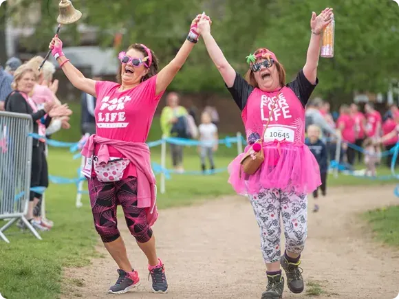Two women in matching pink "Race for Life" t-shirts and festive accessories run along a path during a charity race event, with spectators and barriers lining the route.