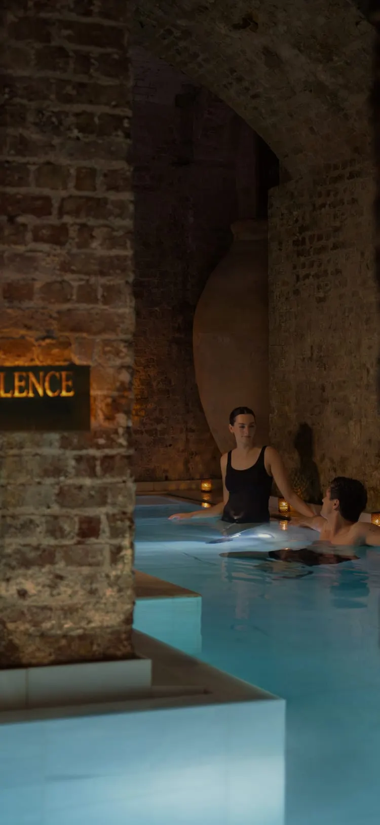 Two people bathing in luminous turquoise thermal water within a historic stone-walled underground chamber lit by candlelight.