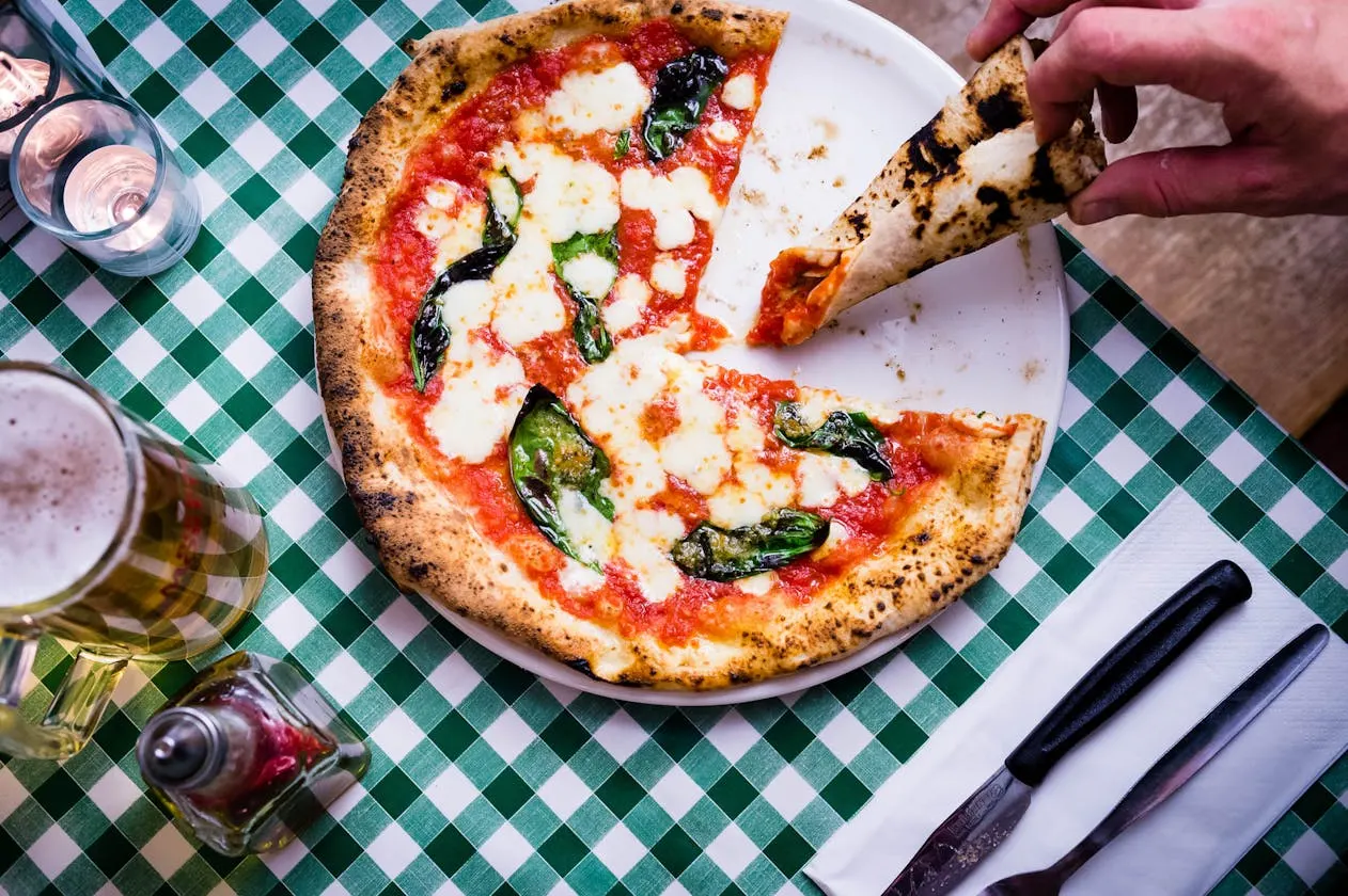 Person picking up a slice of tomato and mozzarella pizza with basil on a checked tablecloth.